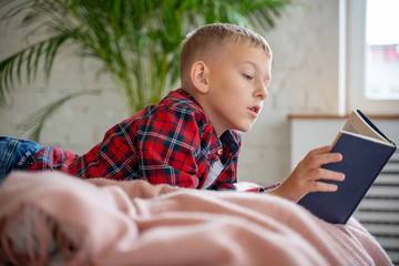 Boy, a schoolboy of 8 years old, reads with interest a paper book on the bed in the bedroom, education, children's literature, development of imagination
