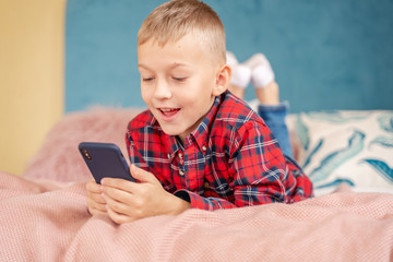 Happy joyful child 8 years old schoolboy lies on bed with mobile phone in his hands, education game social media concept