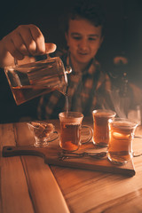 a young man pours tea from a hot teapot into a clear Cup on a wooden table in the evening on dark background