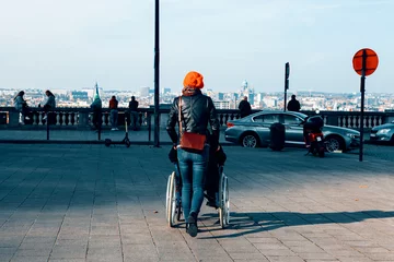 Rolgordijnen rear view shot of a woman pushing a wheelchair with a patient sitting on it with the metropolitan city in background - health care and disability concept © Carlo