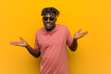 Young casual black man wearing sunglasses doubting and shrugging him shoulders in questioning...