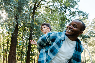 low angle view of happy african american piggybacking cute curly son near soap bubbles in park