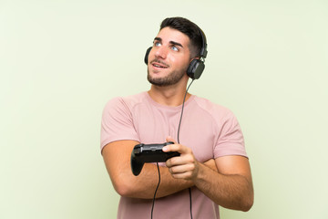 Young handsome man playing with a video game controller over isolated green wall looking up while smiling
