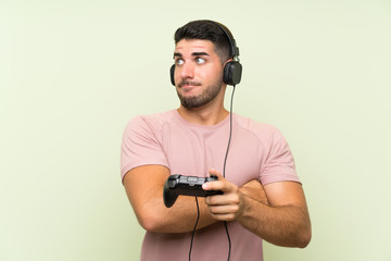Young handsome man playing with a video game controller over isolated green wall making doubts gesture while lifting the shoulders © luismolinero