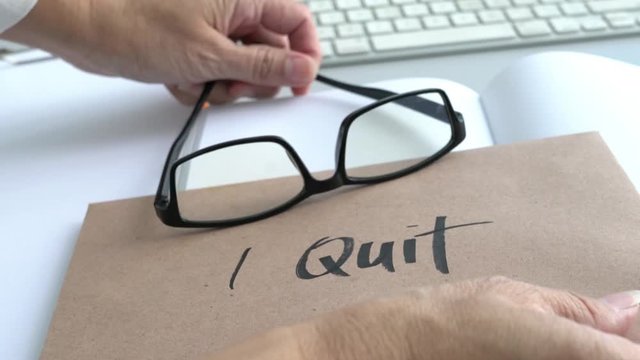 Putting a brown envelope with I Quit words with reading glasses on top of it, on an office desk with notepad . Resignation or quit job concept.