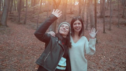 Two beautiful girl is waving hand in a forest, autumn time. Happy woman, greeting and saying hi with gestures. 