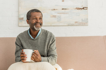 senior, positive african american man sitting with blanket on knees and holding cup of warming drink