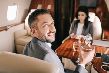 selective focus of happy businessman holding jar with alcohol drink near businesswoman in private jet