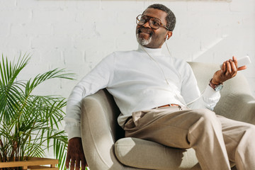 stylish african american man sitting in armchair and listening music in earphones