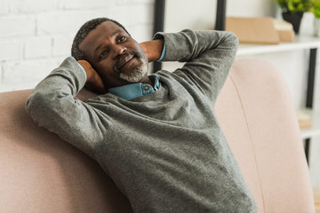 dreamy, positive african american man looking away while sitting on sofa