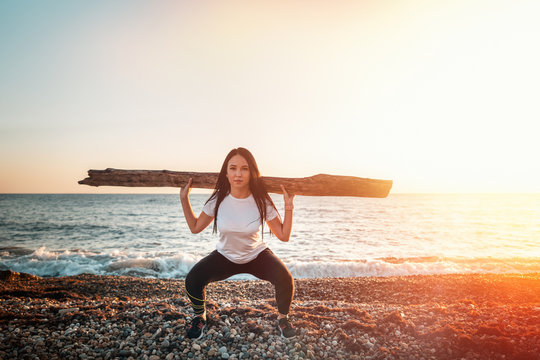 Concept of sport and healthy lifestyle. A woman in sportswear raises a log, engaged in strength exercises in the fresh air. In the background the sea and the sky. Copy space. Light