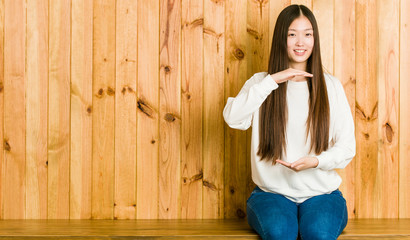 Fototapeta premium Young chinese woman sitting on a wooden place holding something with both hands, product presentation.