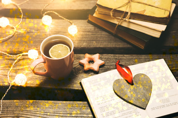 Holidays mood photo. Christmas lights and hot tea mug. Book for cosy evening. Sweet gingerbread and wooden heart on tray. Perfect winter flat lay. Hygge concept.