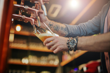 Tattooed caucasian barman pouring beer while standing in pub. Selective focus on hand.