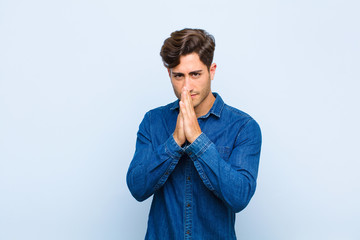 young handsome man feeling worried, hopeful and religious, praying faithfully with palms pressed,...