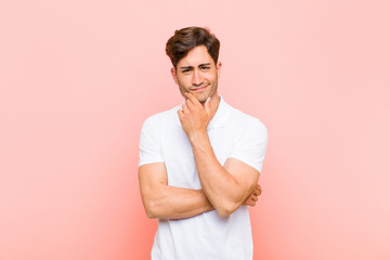 young handsome man looking serious, thoughtful and distrustful, with one arm crossed and hand on...