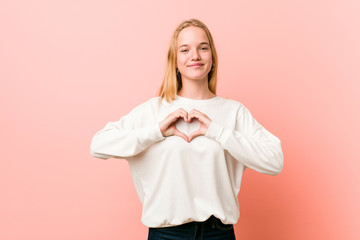 Young blonde teenager woman smiling and showing a heart shape with hands.
