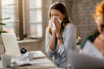 Young businesswoman using a tissue while sneezing in the office. - obrazy, fototapety, plakaty