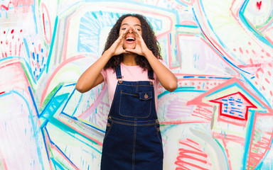 young pretty african american woman feeling happy, excited and positive, giving a big shout out with hands next to mouth, calling out against graffiti wall