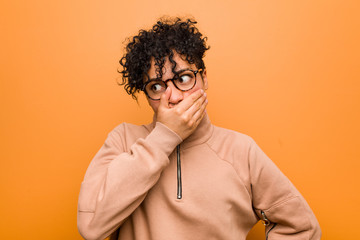 Young mixed african american woman against a brown background thoughtful looking to a copy space covering mouth with hand.