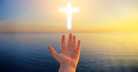 Human hand open palm pray to Christian cross light above sunset water. Jesus Christ church faith...