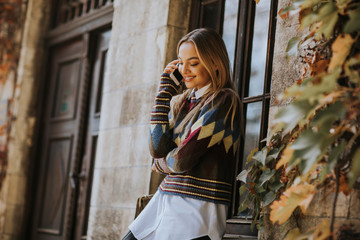 Pretty young woman standing with mobile phone on street at autumn day