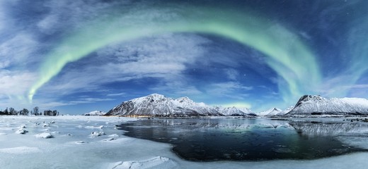 Wide shot of the arch shaped northern lights over a frozen sea with the snow covered mountains