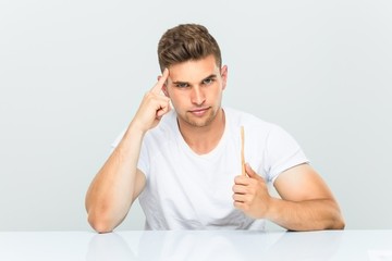 Young man holding a toothbrush pointing his temple with finger, thinking, focused on a task.