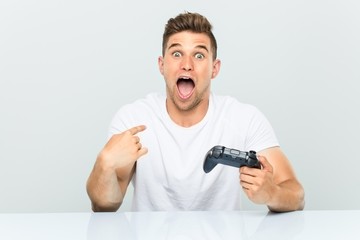 Young man holding a game controller surprised pointing at himself, smiling broadly. © Asier