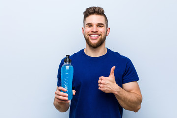 Young caucasian sportsman holding an isotonic drink smiling and raising thumb up
