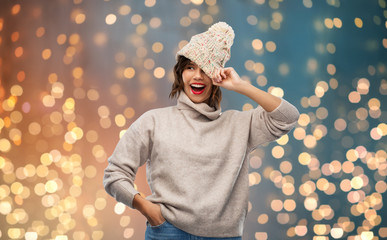 christmas, season and people concept - happy smiling young woman in knitted winter hat and sweater over festive lights background