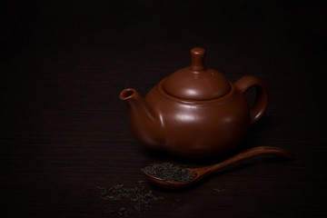 Clay teapot on a dark wooden table. Nearby are tea leaves in a wooden spoon.