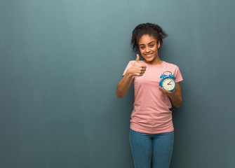 Young black woman smiling and raising thumb up. She is holding an alarm clock.