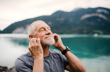 Senior man with earphones standing by lake in nature, listening to music.