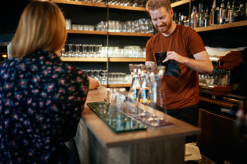 Handsome caucasian barman staning in bar and wiping pint and talking to customers. Customers drinking beer. Nightlife.