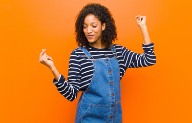 young pretty black woman smiling, feeling carefree, relaxed and happy, dancing and listening to...