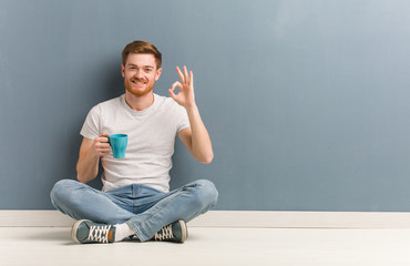 Young redhead student man sitting on the floor cheerful and confident doing ok gesture. He is holding a coffee mug.