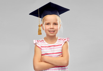 school, education and learning concept - happy little girl in bachelor hat or mortarboard over grey background