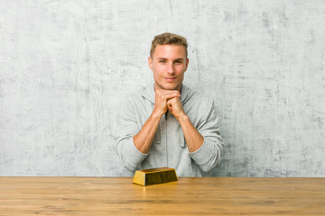 Obraz premium Young handsome man holding a gold ingot on a table keeps hands under chin, is looking happily aside.