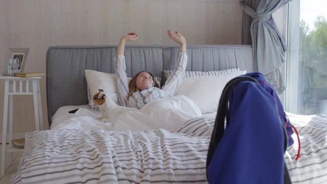 Tracking of happy woman in pajama waking up in morning and stretching in bed, then petting cute dog lying beside her. Blue jiu-jitsu gi with black belt hanging on chair
