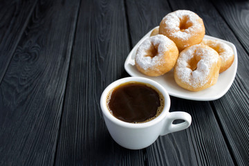 Black coffee in the white cup and homemade donut with powdered sugar on the black wooden background. Copy space. Closeup.