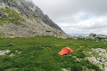 Red and orange tent pitched on a green meadow in high alpine mountain landscape. Outdoor overnight sleeping in a tent. Green and yellow meadow with flowers and a trekking tent. Image shot of a tent.