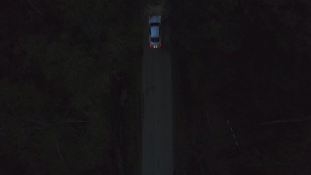Aerial top view silver premium car Cadillac Cts driving on dark countryside road among summer forest at night. View from drone white car with xenon headlights riding on night road through dark forest