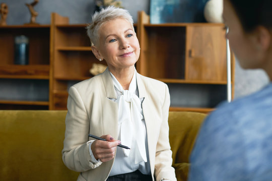 Senior Company Executive With Short Stylish Haircut Sitting In Office With Pen Smiling While Listening To Experience Of Job Candidate During Interview. Mature Female Interviewer Recruiting Assistant