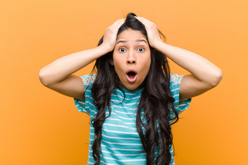 young pretty hispanic woman feeling horrified and shocked, raising hands to head and panicking at a...