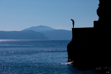Tourists jumping off a large rock ledge in Amoudi Bay on Santorini Island in Greece.