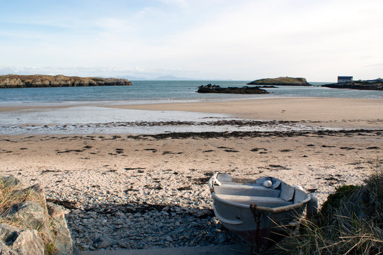 A clear, crisp spring day at the seaside on the Welsh island of Anglesey.  Bright sunlight. A solitary row boat at the edge of the sand dunes.  Low tide with sand flats. An unspoiled, pristine beach.