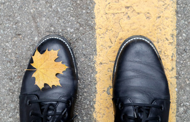A beautiful combination of a yellow maple autumn leaf next to the yellow road markings and the legs of a girl in black boots in the park. Autumn concept, beautiful background with copy space