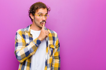 young handsome man asking for silence and quiet, gesturing with finger in front of mouth, saying...