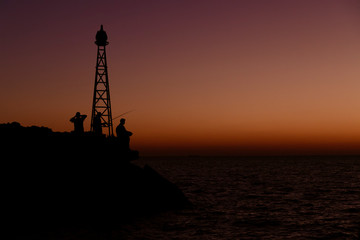 Obraz premium Fishermen standing on rocks near a light beacon in front of a vibrant sunset on Santorini Island in Greece.